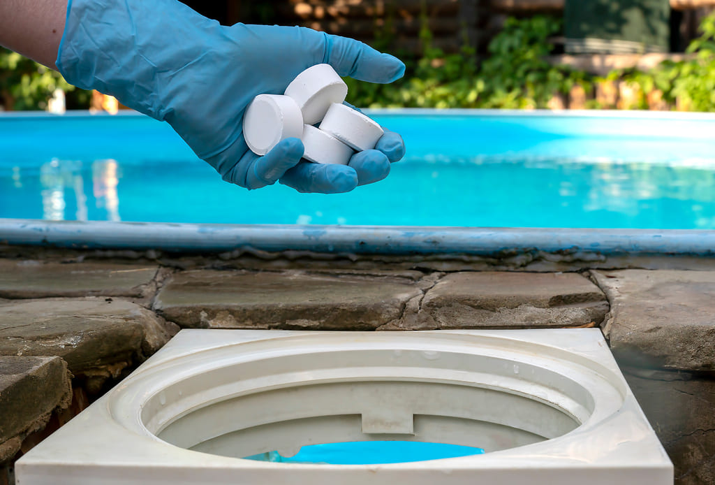 Main avec un gant bleu déposant des galets de chlore dans le skimmer de la piscine